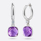 An angled view of two silver drop hoop earrings, showing the faceted surface of the cushion-cut violet amethyst centrepieces.
