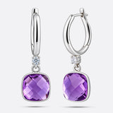 An angled view of two silver drop hoop earrings, showing the faceted surface of the cushion-cut violet amethyst centrepieces.