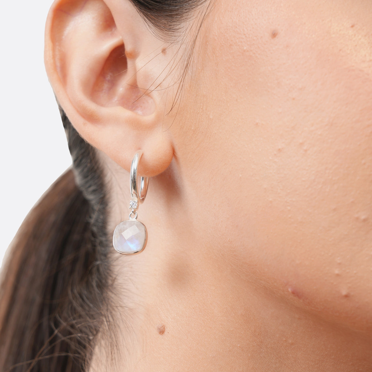 Close-up of a silver drop hoop earring worn by a model, showing the cushion-shaped Rainbow Moonstone and its blue iridescence.